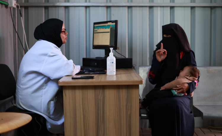 A healthcare worker in a white coat, black head scarf and glasses sits at a desk in front of a computer, listening to a woman in all black clothing who is holding a baby