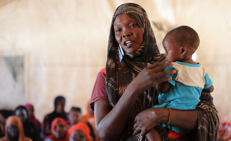 A woman in a green and orange head scarf stands holding a baby in a blue jumpsuit; other women seated behind her are visible in the background