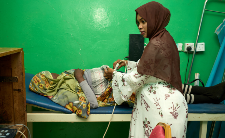 A woman in a brown headscarf and white dress with flower patterns hold an ultrasound scanner over the belly of a pregnant woman in a green and yellow headscarf, lying on a blue hospital bed in front of a bright green wall