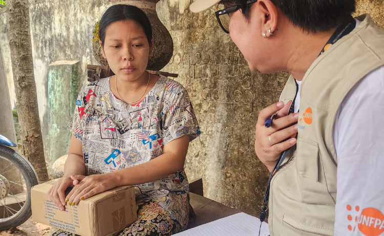  A woman sits on a wooden bench holding a cardboard box, beside another woman in UNFPA-branded clothing, a cap and glasses
