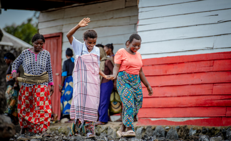 Two women smile and hold hands as they walk along a muddy path outside a red and white painted building. Another woman walks behind them and more are seen waiting outside the building in the background