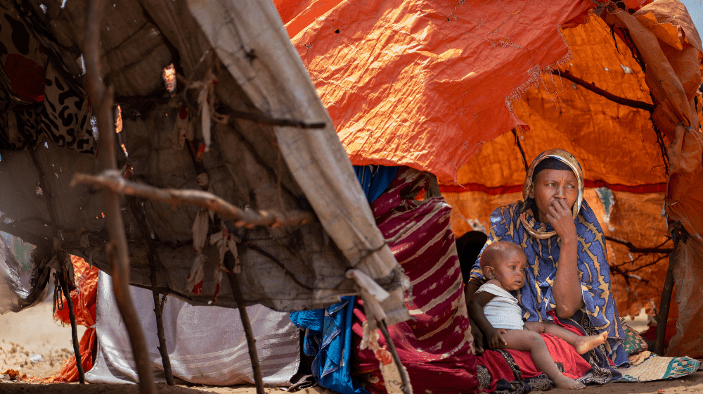 A woman dressed in blue and yellow sits on the ground with a baby on her lap in front of an orange tarpaulin tent